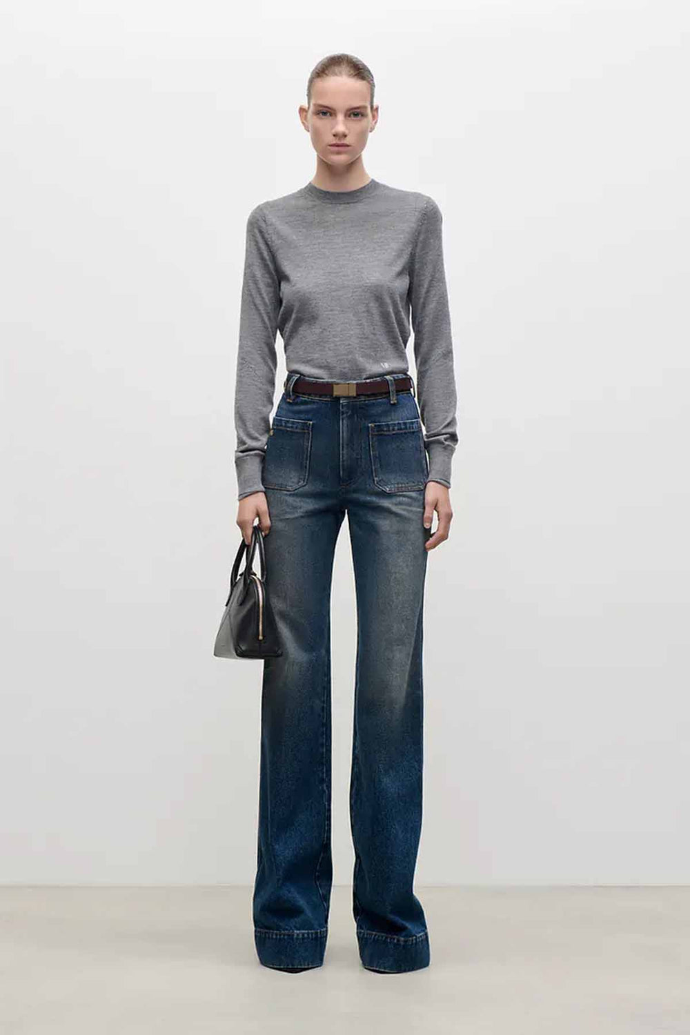 Modell med klær fra Victoria Beckham, jeans med brunt belte, sort liten veske og grå genser. Hos Moniker.